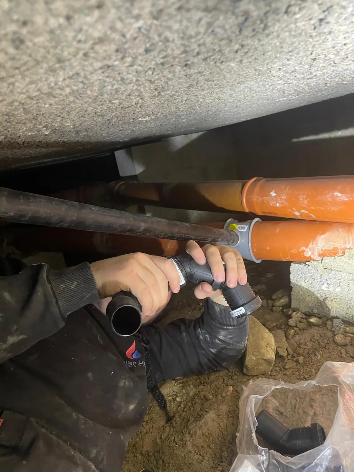 Technicien d'ABL Chauffage préparant un raccord d'évacuation noir sur une canalisation orange, dans un vide sanitaire