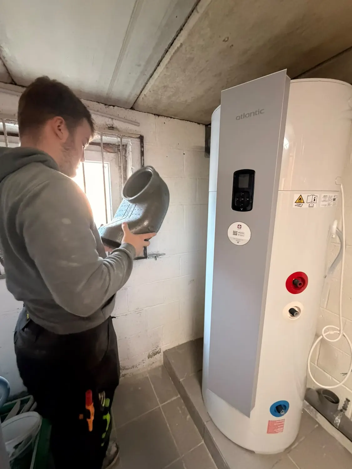Technicien tenant un coude de gaine isolé à côté d'un boiler thermodynamique Atlantic